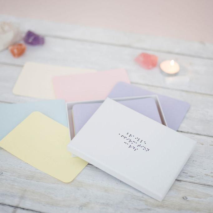 A white box with black braille dots reading braille affirmation cards, there are coloured sheets of paper with braille on, crystals and a candle in the background. 