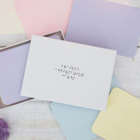 A white box with black braille dots reading braille affirmation cards, there are coloured sheets of paper with braille on.