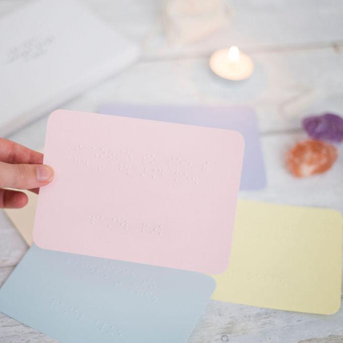 A hand holding a piece of pink card with an affirmation in braille, in the background are more coloured pieces of card, crystals and a candle.