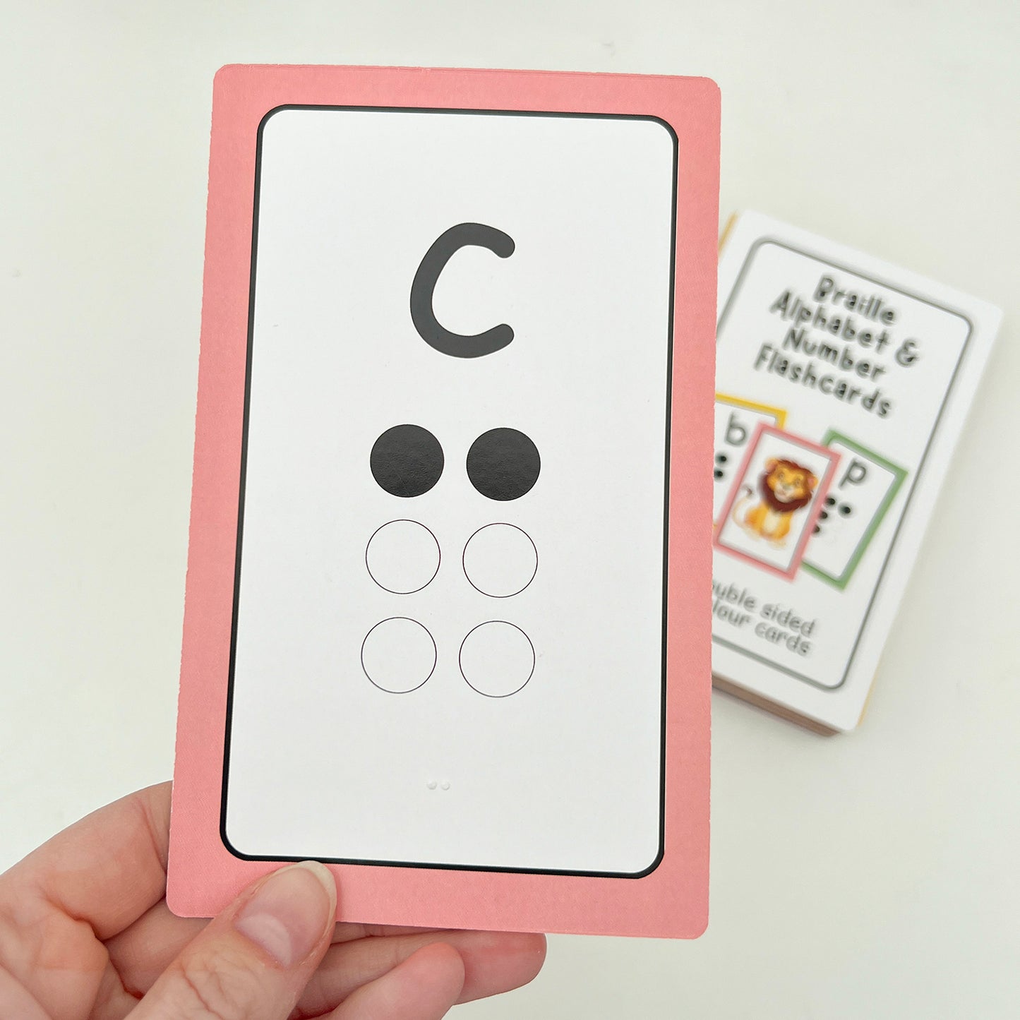 A hand holding a flashcard with the letter C in print, printed braille dots and tactile braille dots. The rest of the pack are in the background.