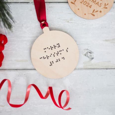 A round wooden Christmas tree decoration with red ribbon and braille that reads Merry Christmas 2024.