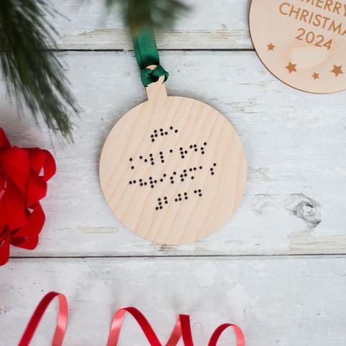 A round wooden Christmas tree decoration with green ribbon and braille that reads The Kellard Christmas 2024.