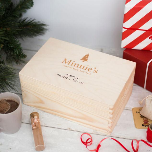 A wooden Christmas Eve Box with Minnie's Christmas Eve Box in print and lower case grade one braille. There are Christmas gifts visible in the top right of the image, a Christmas tree in the top left and a hot chocolate in the bottom left.