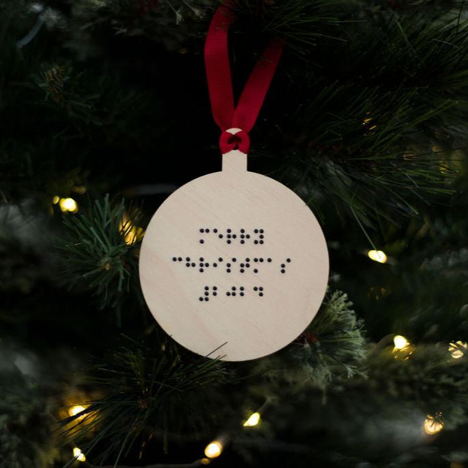 A round wooden Christmas tree decoration with Merry Christmas 2024 in lower case braille. The bauble has red ribbon and is hanging on a Christmas tree.