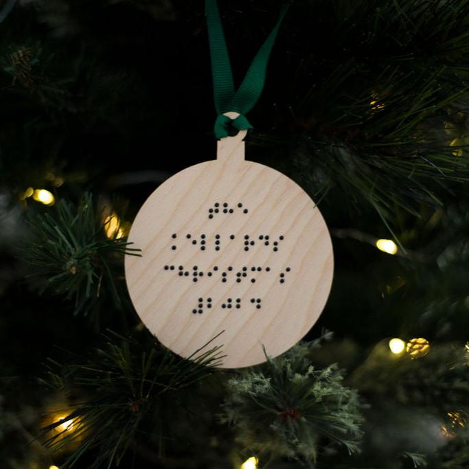 A round wooden Christmas tree decoration with The Kellard Christmas 2024 in lower case braille. The bauble has green ribbon and is hanging on a Christmas tree.