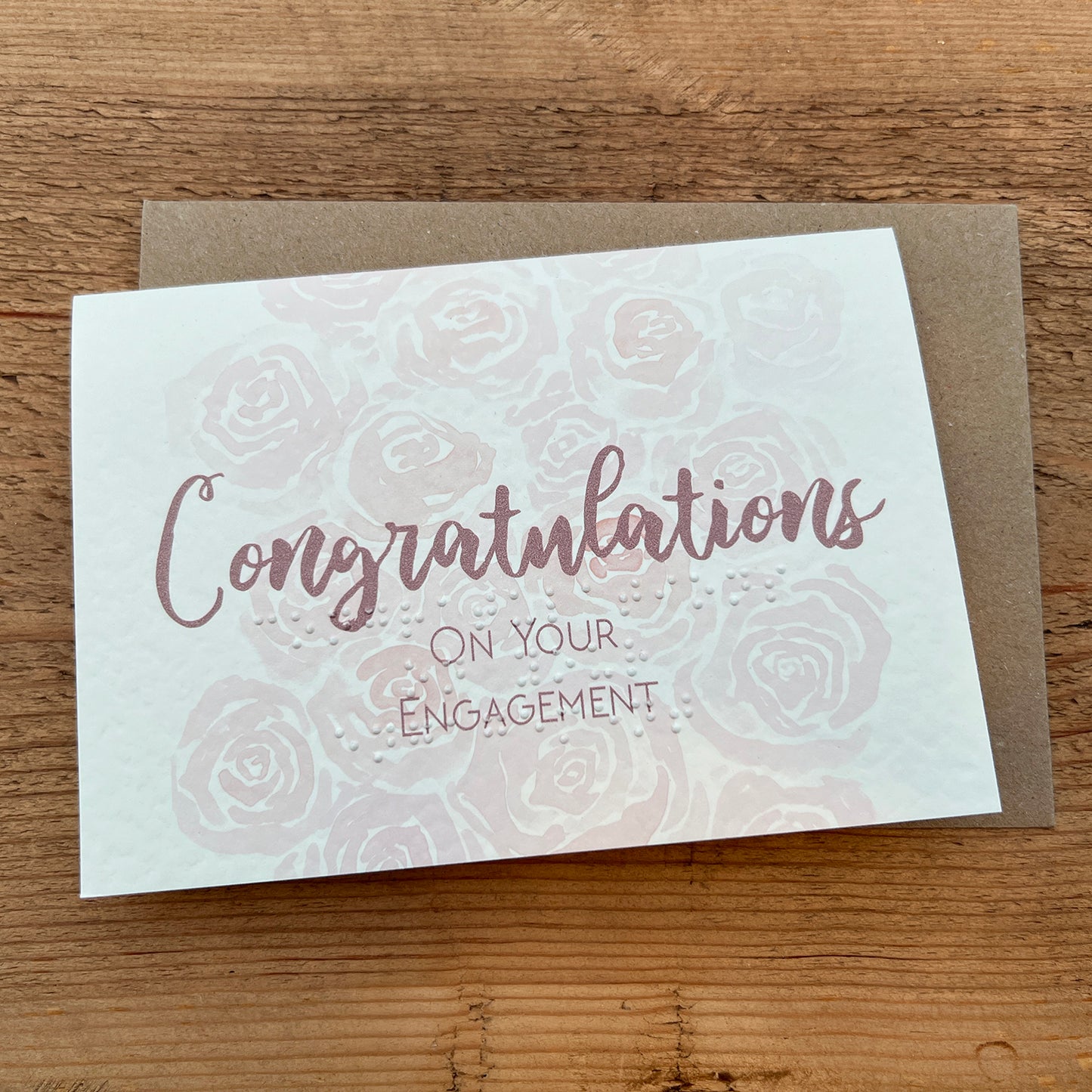 A greeting card that reads "Congratulations on Your Engagement" in a stylish, cursive font, this is also typed in braille underneath. The background of the card is adorned with a subtle pattern of roses, giving it a delicate and celebratory feel.