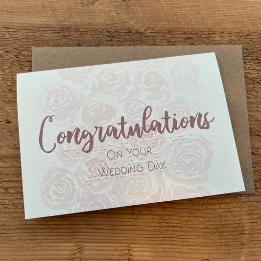 A greeting card that reads "Congratulations on Your Wedding Day" in a stylish, cursive font, this is also typed in braille underneath. The background of the card is adorned with a subtle pattern of roses, giving it a delicate and celebratory feel.