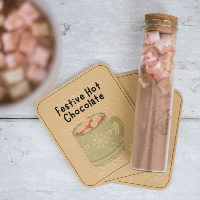 A glass tube of hot chocolate and marshmallows, with a mug of hot chocolate and marshmallows, and a label saying festive hot chocolate in both print and braille.