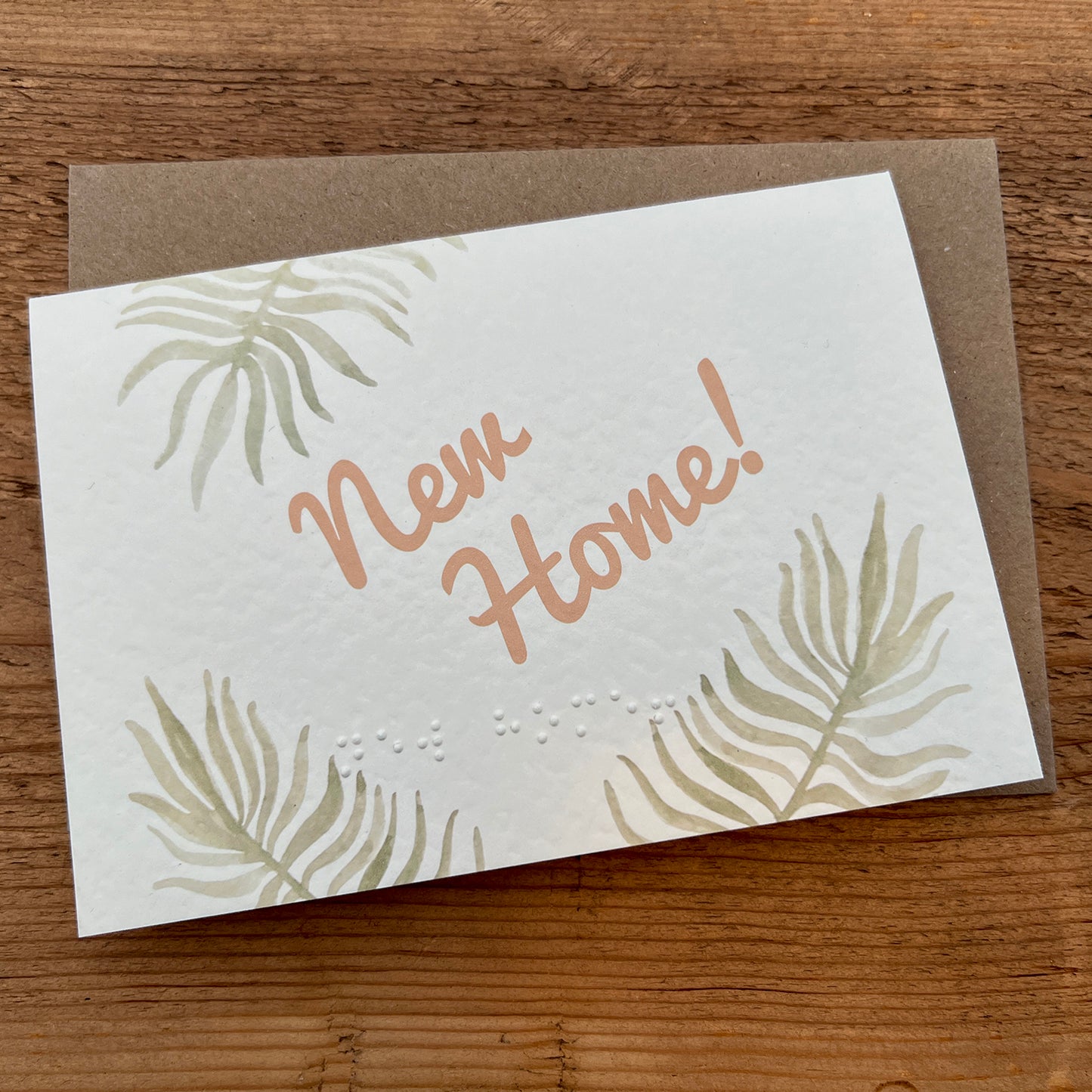 A greeting card with the words "New Home!" written in a stylish, cursive font, the same message is also typed in braille beneath it. The background of the card features a soft, leafy design with light green palm leaves in the corners.