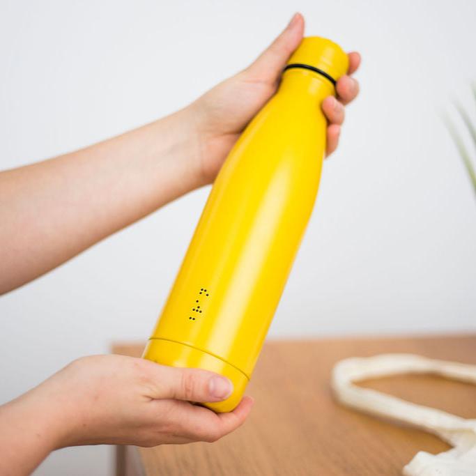 Hands holding a yellow water bottle with bran written in lower case black braille dots.