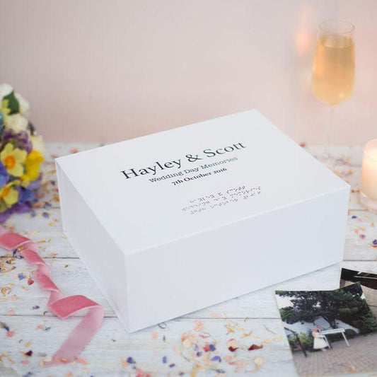 A large white memory box with black text reading Hayley & Scott, Wedding Day Memories, 7th October 2016 in both print and braille. There is a bridal bouquet in the top left, a candle and champagne in the top right and wedding photos in the bottom right.