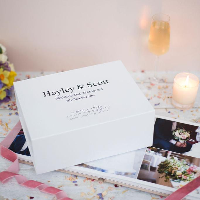 A large white memory box with black text reading Hayley & Scott, Wedding Day Memories, 7th October 2016 in both print and braille. There is a bridal bouquet in the top left, a candle and champagne in the top right and wedding photos in the bottom right.