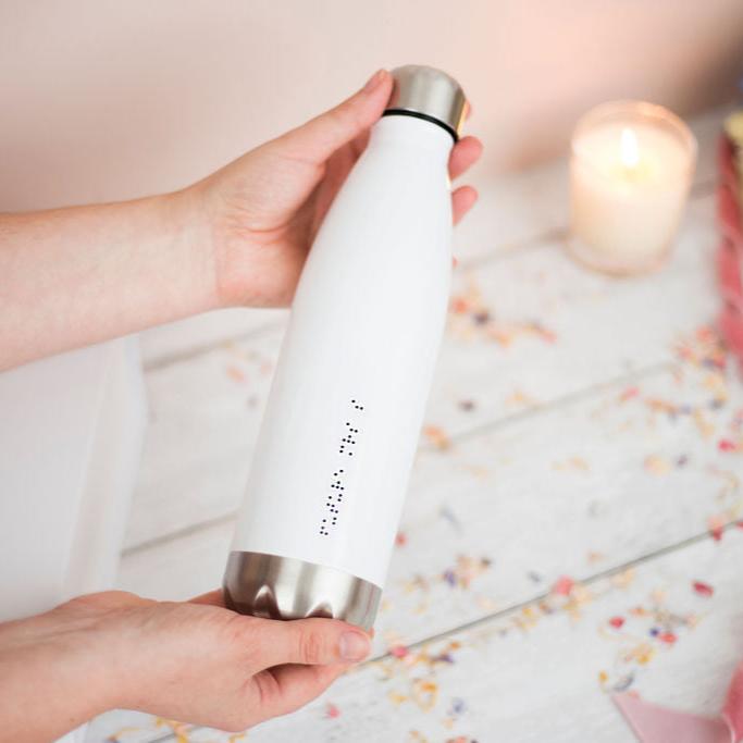 Hands holding a glossy white water bottle, with future Mrs S in braille, the dots are black and raised.