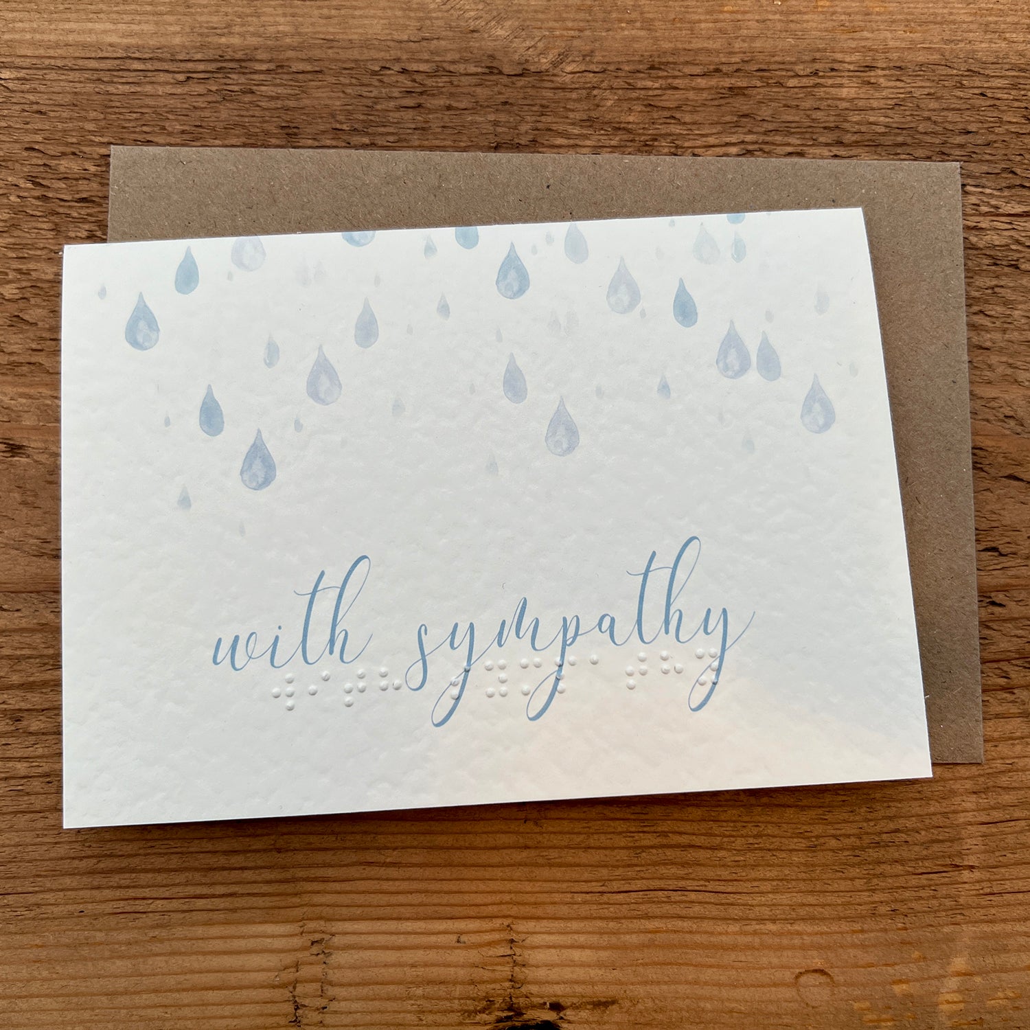 A greeting card with the words "with sympathy" written in a gentle, cursive font, the same message is also typed in braille beneath it. The background of the card features a simple design of soft, blue raindrops falling from the top.