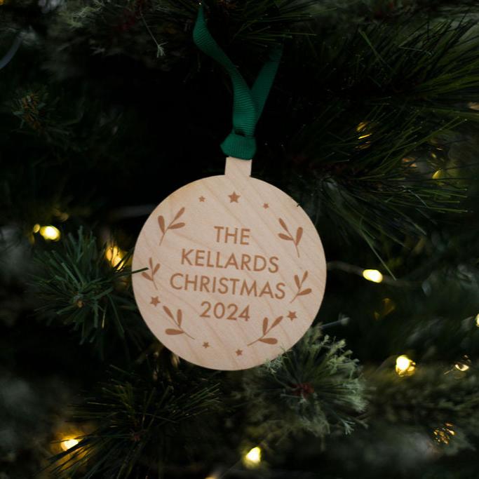 A round wooden Christmas tree decoration with The Kellard Christmas 2024 in printed text and leaf patterns dotted around the edge . The bauble has green ribbon and is hanging on a Christmas tree.