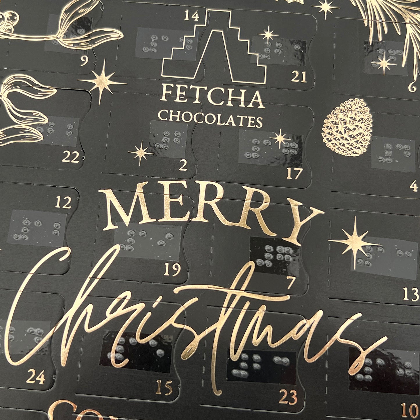 A close up of the advent calendar, a black box with gold writing, you can several of the doors which have both printed text and braille. Other text says Fetcha Chocolates, Merry Christmas.