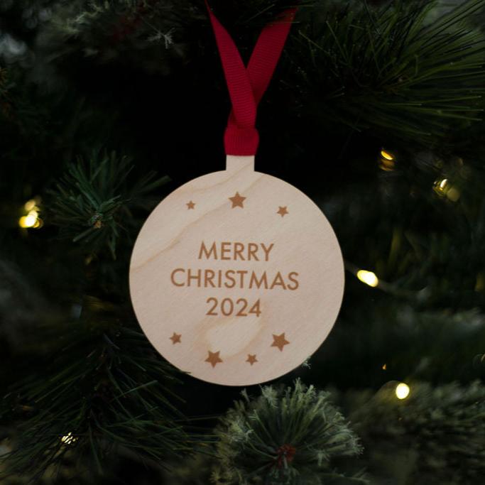 A round wooden Christmas tree decoration with Merry Christmas 2024 in printed text and stars dotted around the edge . The bauble has red ribbon and is hanging on a Christmas tree.