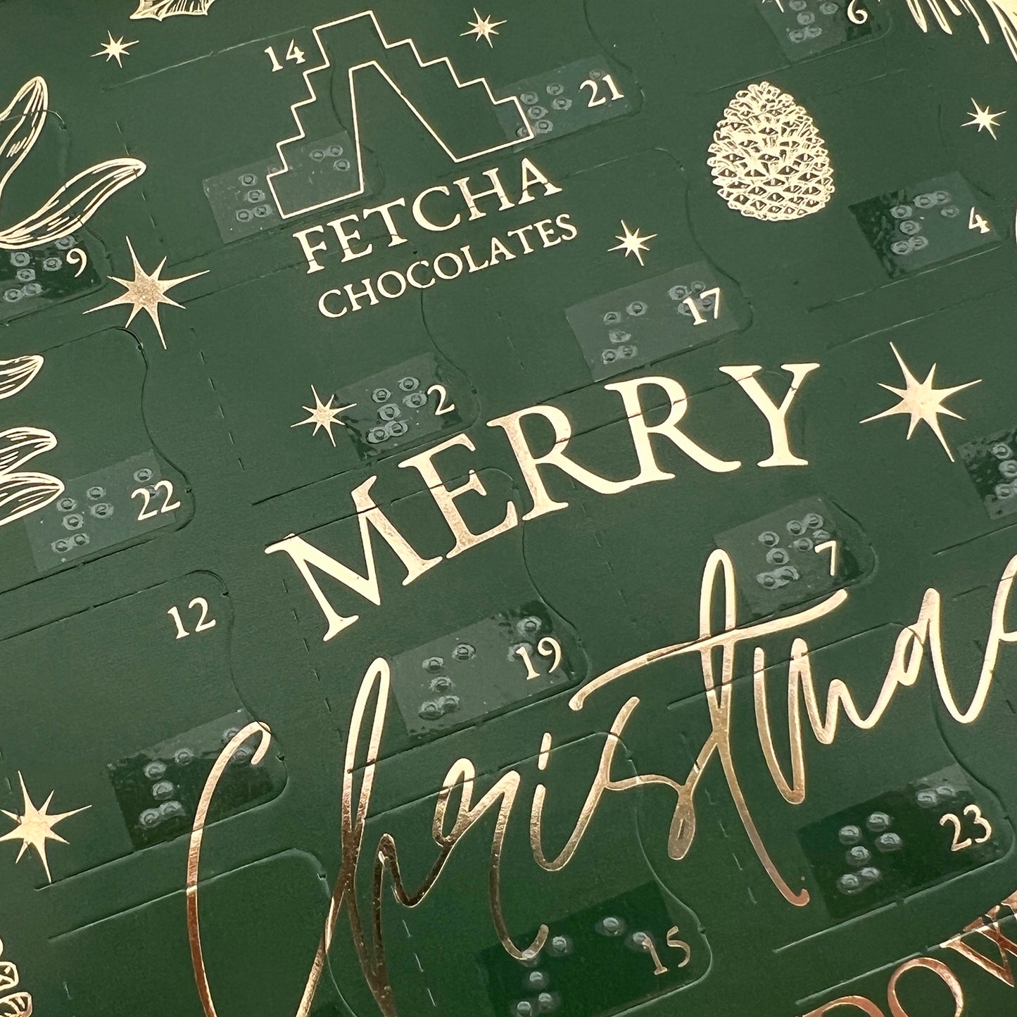 A close up of the advent calendar, a green box with gold writing, you can several of the doors which have both printed text and braille. Other text says Fetcha Chocolates, Merry Christmas.