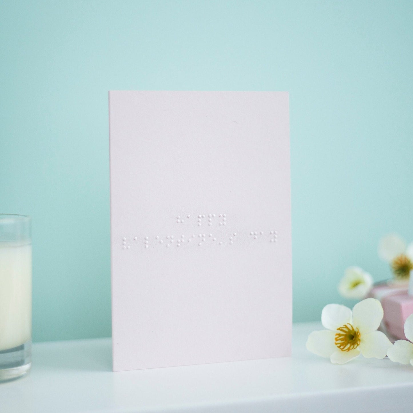 A pastel pink card with Happy Valentine&#39;s Day written in lower case braille. There is a candle to the left and a flowers to the right.